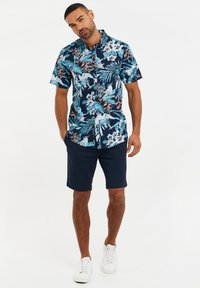 Korte mouwen marineblauwe bloes met felle blauwe, groene en oranje tropische patronen, gecombineerd met donker marineblauwe shorts en witte sneakers.