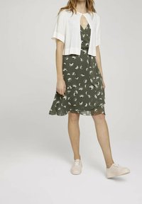 Abito floreale verde con orlo a strati, abbinato a un cardigan corto bianco. La modella indossa sneakers di colore chiaro. Tessuto dalla consistenza liscia.