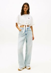 Camiseta blanca recortada con un detalle de nudo en el frente, combinada con jeans de pierna ancha en un lavado claro. Zapatillas completan el atuendo casual.