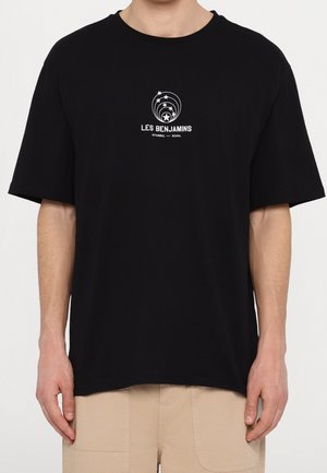 Czarny luźny t-shirt z białym logo "Les Benjamins Istanbul—Seoul" z pięcioma gwiazdkami nad tekstem, noszony z beżowymi spodniami.