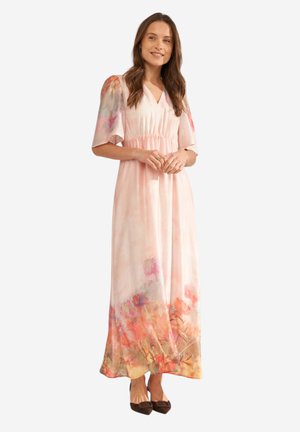 Kobieta stojąca, ubrana w długą, jasnoróżową sukienkę w kwiaty z krótkimi rękawami i brązowe, zaostrzone buty, uśmiechająca się z złączonymi rękami.