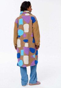 Kobieta widziana z tyłu, ubrana w długi beżowy płaszcz z dużymi niebieskimi, fioletowymi i białymi abstrakcyjnymi łatami, niebieskie dżinsy i różowe buty.