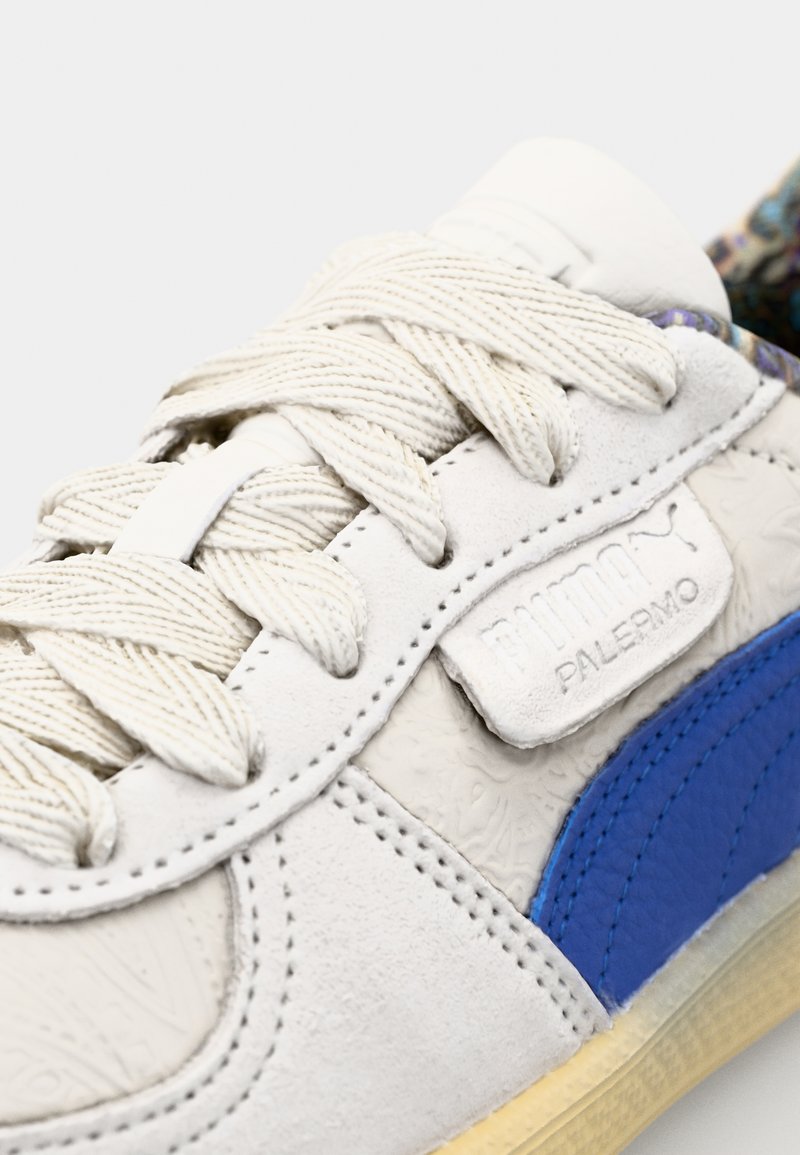 La zapatilla Puma Palermo presenta un upper de ante blanco texturizado, un panel lateral de cuero azul y agujetas crema con una etiqueta de logo en la lengüeta.