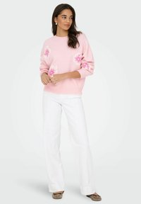 Pull en tricot rose avec des motifs floraux, col rond et coupe décontractée, associé à un pantalon blanc à jambes larges et des ballerines marron.