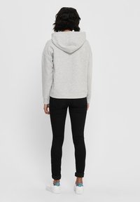Felpa grigia con cappuccio dal morbido texture, abbinata a jeans neri attillati e sneakers bianche con un accento metallico sul tallone.