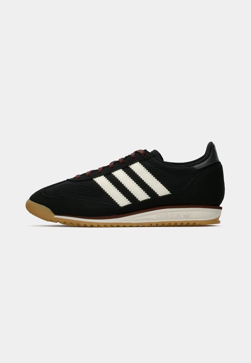Zapatilla deportiva negra con parte superior texturizada, que presenta tres franjas blancas en el lateral, una suela de goma marrón y cordones de color burdeos oscuro.
