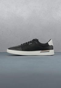 Baskets basses en cuir noir avec semelle et talon blancs, lacets noirs, et logo "BOSS" sur le côté, montrées en profil latéral sur une surface grise.