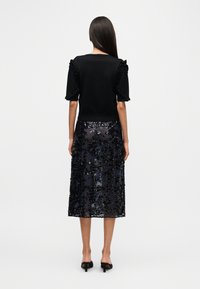 Μαύρο τοπ με βολάν στα μανίκια, συνδυασμένο με διάφανο, μαύρο φούστα με παγιέτες. Το σύνολο έχει εφαρμοστή σιλουέτα και μήκος μέχρι το μέσο της γάμπας.