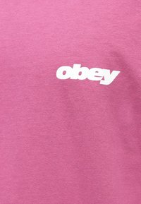 Růžové bavlněné tričko s bílým logem "obey" v moderním písmu. Materiál má jemnou, hladkou texturu a uvolněný střih.