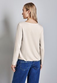 Vaalean beige pitkähihaista puseroa, jossa on rento malli, pyöreä pääntie ja sileä pinta, yhdistettynä sinisiin farkkuihin, joissa on takataskut.