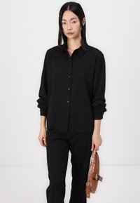 Black long-sleeve button-up shirt with a collar, relaxed fit, and rolled cuffs. Model holds a brown crocodile-embossed handbag.