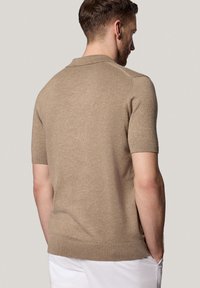 Beżowa koszulka polo z krótkim rękawem, wykonana z dzianiny, z prążkowanym kołnierzykiem i mankietami, dopasowanym krojem oraz gładką teksturą. Brak widocznych logo.