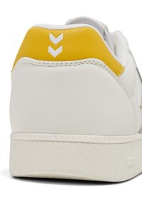 White sneaker with a textured rubber sole, yellow heel accent, and white chevron detail. Smooth leather upper with contrasting stitching.