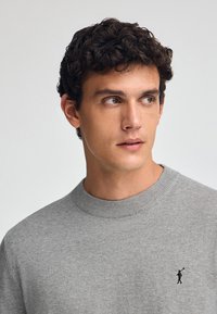 Maglione grigio a maglia con scollo rotondo a coste e piccolo logo scuro sul petto. Tessuto liscio con una vestibilità classica.
