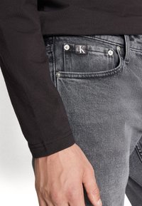 Chemise noire à manches longues associée à un jean gris foncé avec un effet délavé, des accessoires en métal argenté et une étiquette avec le logo CK sur la poche avant.