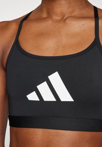 Svart sports-bh laget av glatt stoff, med et hvitt geometrisk logodesign på forsiden. Tynne stropper og en tettsittende passform.