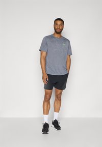 Grijze sportt-shirt met groen logo, gecombineerd met zwarte gestructureerde shorts. Gemaakt met zwarte sneakers en witte sokken, staande op een neutrale achtergrond.