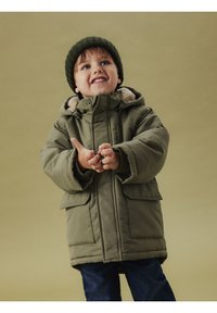 Oliwkowa puchowa kurtka z dwoma przednimi kieszeniami, kapturem wyściełanym sztucznym futerkiem oraz dzianinową czapką. Noszona z niebieskimi dżinsami.