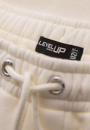 Tessuto color crema con una finitura testurizzata. Presenta un cordino con occhielli in metallo argentato e un'etichetta nera stampata con "LEVEL UP."