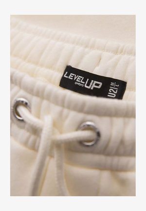 Krämfärgad tyg med en strukturerad yta. Har en dragsko med silverfärgade metallöglor och en svart etikett präglad med "LEVEL UP."