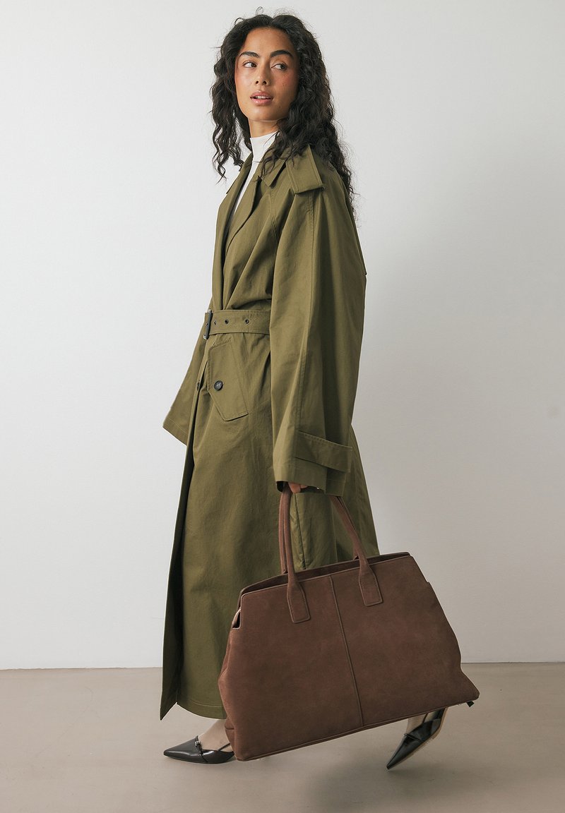 Olive zaļš trenci ar jostas vidu un pogu detaļām, apvienots ar brūnu zamša rokassomu un melnām adītām kurpēm ar norādīto purngalu.
