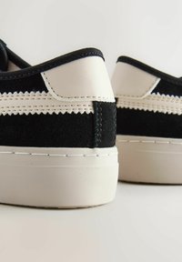 Mustat mokkasneakersit, joissa on valkoinen nahkasaapas ja ompeluyksityiskohtia. Paksu valkoinen kumipohja. Pyöristetty muoto, tyylikäs muotoilu.