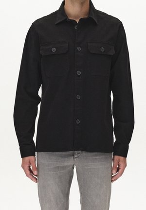 Mann trägt ein langärmeliges schwarzes Hemd mit Knöpfen und zwei Brusttaschen sowie hellgraue Jeans, steht vor einem weißen Hintergrund.