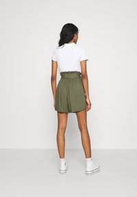 Femme aux longs cheveux noirs portant un haut blanc à manches courtes, un short taille haute vert olive et des baskets montantes blanches, se tenant contre un mur blanc.