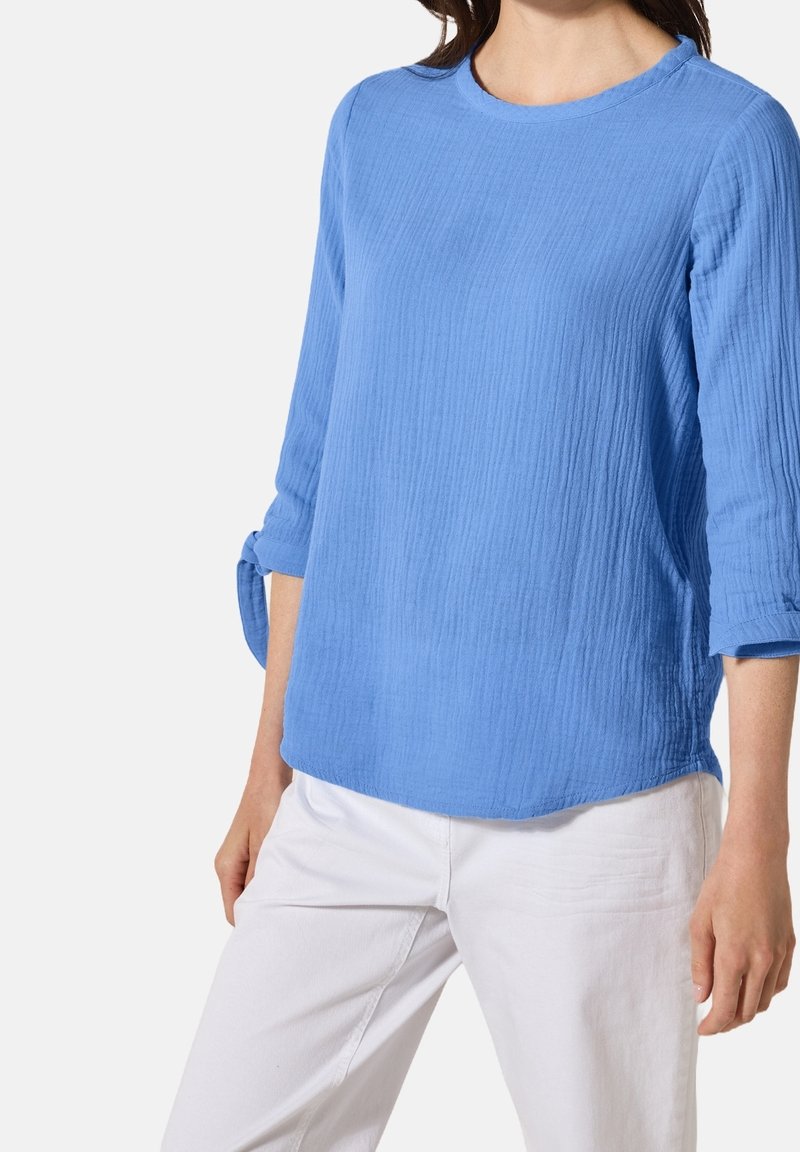 Chemise bleu clair texturée avec un col rond et des manches trois-quarts, présentant des poignets retroussés et une coupe décontractée. Portée avec un pantalon blanc.