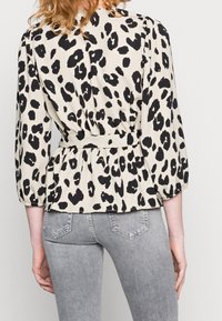 Blusa beige con una stampa leopardata nera, caratterizzata da una vita arricciata, maniche a sbuffo e una texture liscia. Abbinata a jeans grigi.