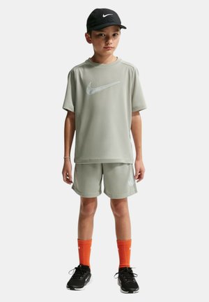 Niño de pie vistiendo camiseta y pantalones cortos Nike gris claro, gorra negra Nike, calcetines naranja Nike y zapatillas negras Nike.
