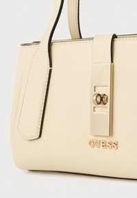 Geantă din piele ecologică crem, cu formă structurată, accesorii aurii și o închidere rectangulară. Prezintă un logo embossat la bază.