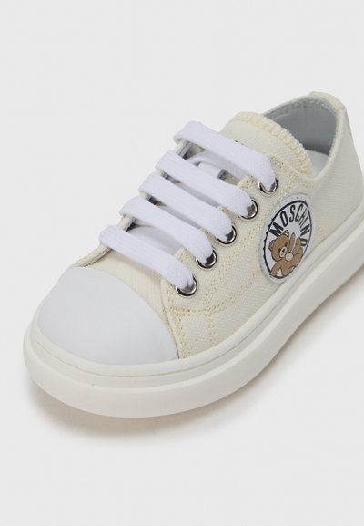 Λευκό πάνινο αθλητικό παπούτσι με γόμα στη μύτη, λευκά κορδόνια και στρογγυλή ετικέτα με λογότυπο που απεικονίζει αρκούδα. Υφή υφάσματος και αντίθετες ραφές.