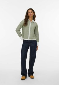 Zielona dziergana kurtka na zamek z białym kołnierzem i mankietami, zestawiona z ciemnoniebieskimi spodniami o szerokich nogawkach i brązowymi butami. Model stoi na tle białego tła.