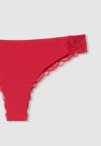 Perizoma rosso realizzato in un tessuto morbido con un delicato bordo in pizzo sulla vita, caratterizzato da un bordo ondulato per un ulteriore dettaglio.