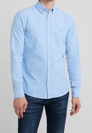 Mann trägt hellblaues Hemd mit Knöpfen und langen Ärmeln mit Markenlogo auf der Brust, kombiniert mit dunkelblauer Jeans, vor schlichtem Hintergrund stehend.
