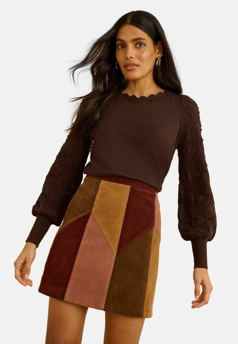 Pull marron avec des manches bouffantes texturées, encolure festonnée ; mini-jupe en daim multicolore avec des blocs géométriques de couleur rouge, marron et beige.