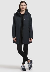 Mörkblå vattentät parka med huva, som har en avsmalnande silhuett, knappslå framtill och dragkedjefickor på sidorna. Bärs över svarta leggings och sneakers.
