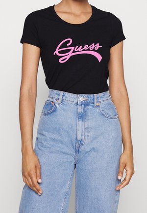Osoba nosząca czarny t-shirt z krótkim rękawem z różowym logo "Guess" oraz jasnoniebieskie jeansy z wysokim stanem, widoczna od szyi do bioder.