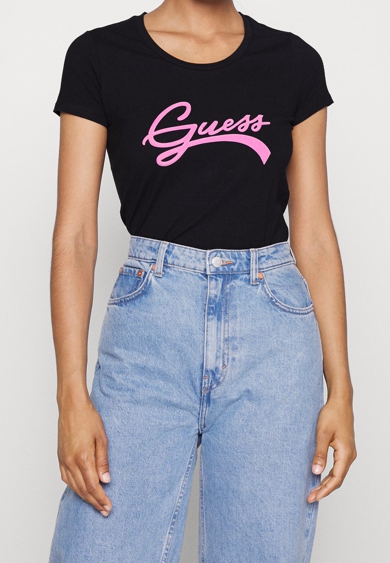 Personne portant un t-shirt noir à manches courtes avec un logo rose "Guess" et un jean taille haute bleu clair, visible du cou jusqu'aux hanches.