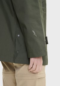 Veste imperméable vert olive avec des manches ajustées, boutons-pression et un logo gris près du poignet. Associée à un pantalon beige clair.