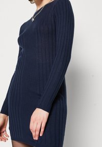 Robe tricotée côtelée navy à manches longues, à coupe ajustée et col rond. Présente une texture subtile et une longueur mi-cuisse.