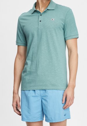 Una camiseta polo verde azulado con un acabado texturizado, que presenta un cierre de tres botones y un pequeño logotipo. Combinada con pantalones cortos azul claro.