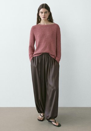 Femme portant un pull en maille rose poudré, un pantalon ample en satin brun foncé et des sandales noires, debout contre un mur clair et uni.