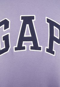 Sudadera de color lavanda con grandes letras en azul marino "GAP" con contorno blanco, presentando una textura de tela suave y un diseño casual.