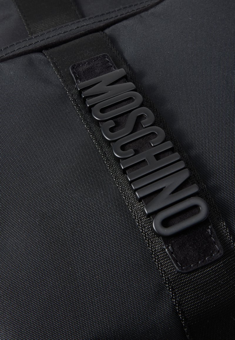 Tissu texturé noir avec une étiquette logo Moschino en caoutchouc noir cousue sur une sangle en tissu noir.