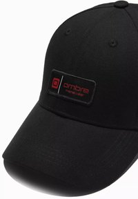 Schwarze Baseballmütze aus Stoff, mit runder Krone, gebogenem Schirm und einem rechteckigen Logo-Patch in Rot und Weiß auf der Vorderseite.