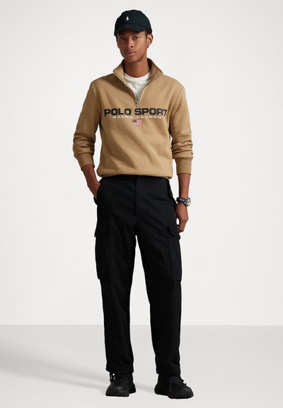 Kollane tõmblukuga dressipluus, millel on "POLO SPORT" logo, koos mustade cargo pükstega. Must müts ja tennised lõpetavad komplekti.