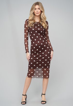 Kvinde med langt blondt hår iført en brun gennemsigtig midikjole med hvide prikker og sorte ankelstroppestiletter, stående foran en ensfarvet grå baggrund.