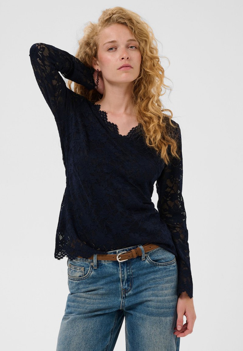 Femme aux cheveux blonds bouclés portant un haut en dentelle noire à manches longues et un jean bleu avec une ceinture marron, posant avec une main derrière la tête.